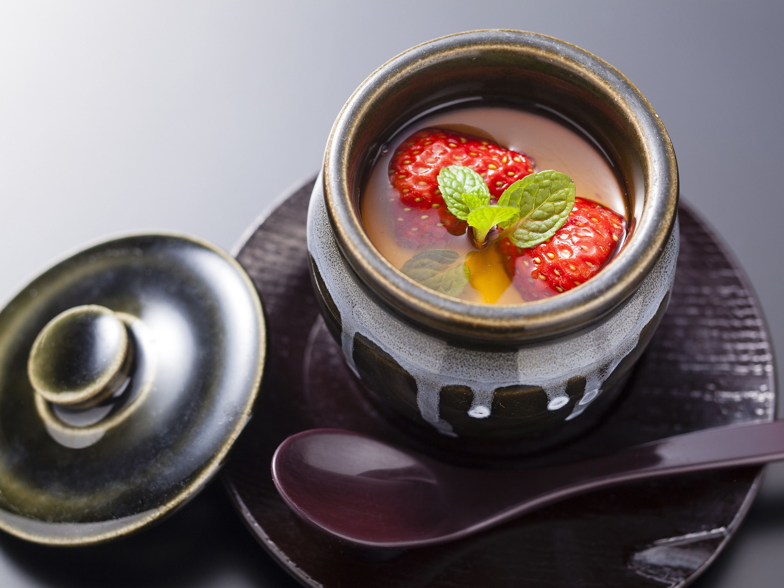 A bowl of soup with strawberries and mint leaves, accompanied by a spoon and a lid.