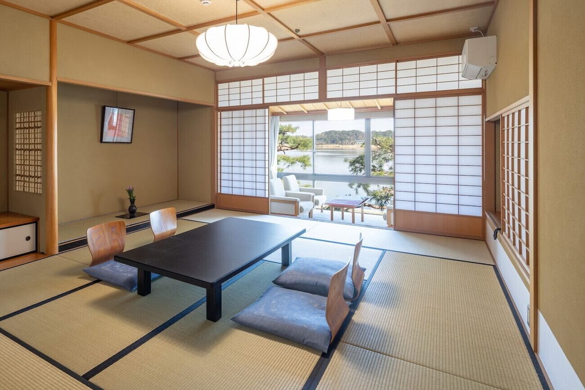 Una habitación con esteras de tatami, una mesa baja y sillas, una lámpara colgante y vistas de un lago a través de biombos shoji.