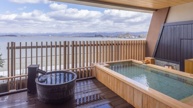 A wooden deck with a hot tub and a stone basin, overlooking a body of water and distant buildings. - APA Hotel & Resort Kaga Katayamazu Onsen Kasuikyo (Kaga)