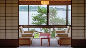 A traditional Japanese room with tatami flooring, a low wooden table, and two chairs. A large window offers a view of a lake and greenery. - APA Hotel & Resort Kaga Katayamazu Onsen Kasuikyo (Kaga)