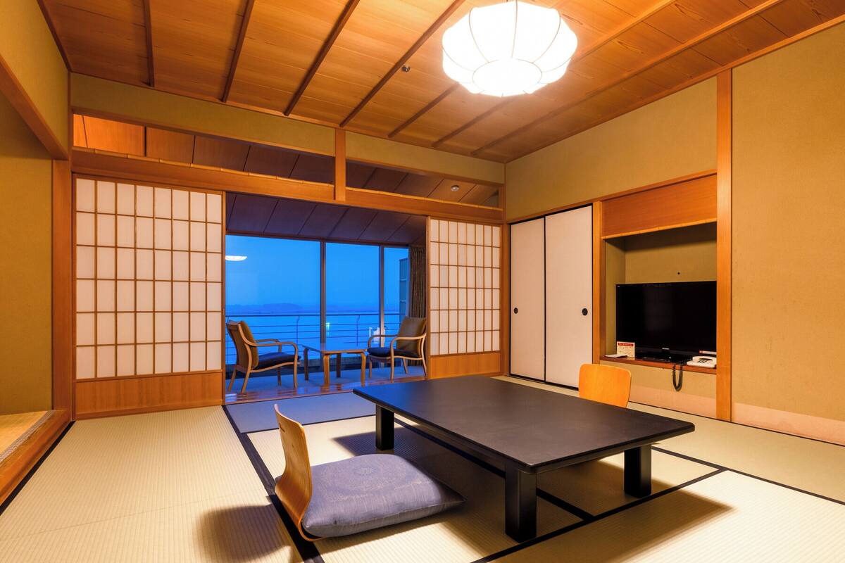 A traditional Japanese room with tatami flooring, a low table, chairs, and a sliding door leading to a balcony with a view.