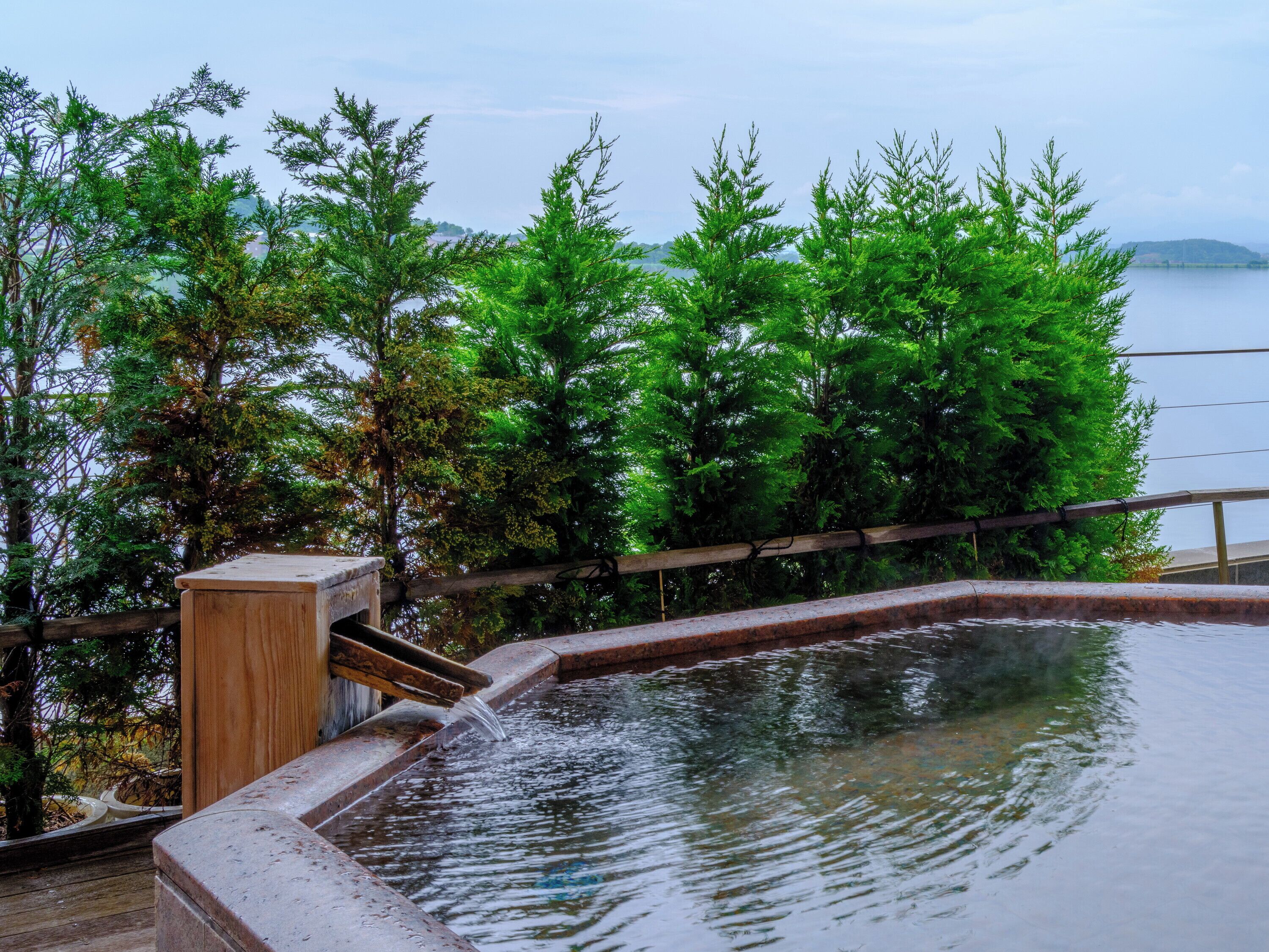 Eine Holzterrasse mit einem kleinen Pool, umgeben von üppiger Vegetation und Blick auf einen See.