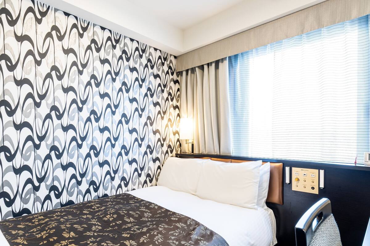 Une chambre d’hôtel avec un mur à motifs, un lit recouvert d’une courtepointe à fleurs, une tête de lit, une table de chevet avec une lampe et une fenêtre avec des rideaux.
