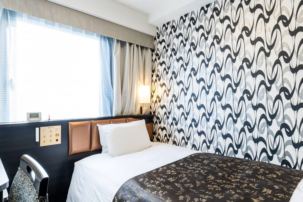 Une chambre d’hôtel comprenant un lit, une tête de lit, une table de chevet, une lampe et un mur recouvert d’un papier peint à motifs.