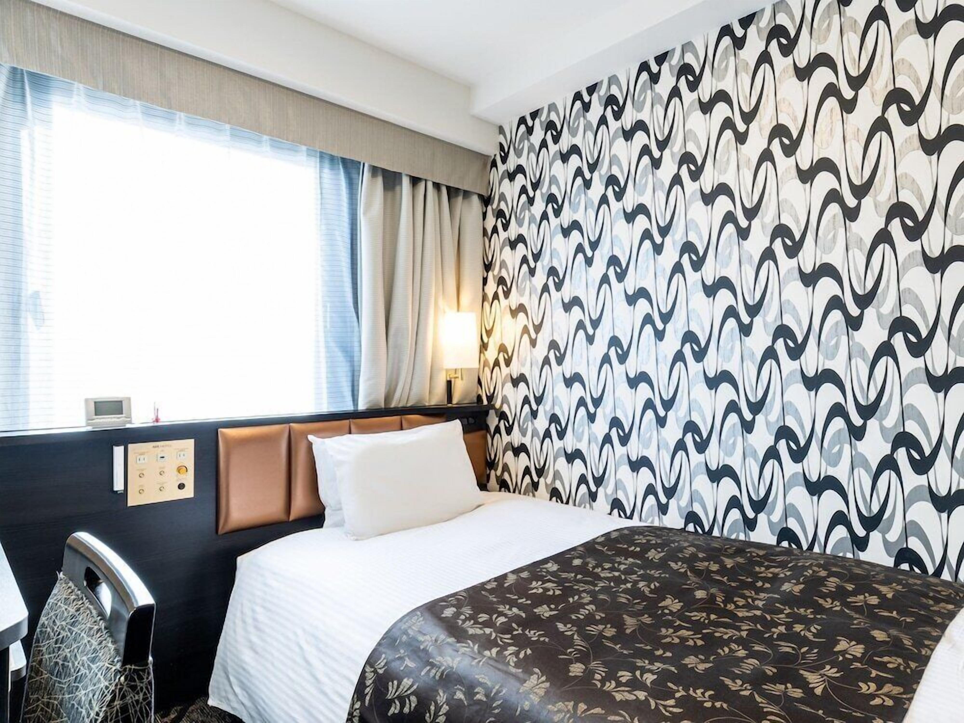 A hotel room with a bed, a desk, and a chair. There is a window with curtains, a wall with a patterned wallpaper, and a wall-mounted control panel.