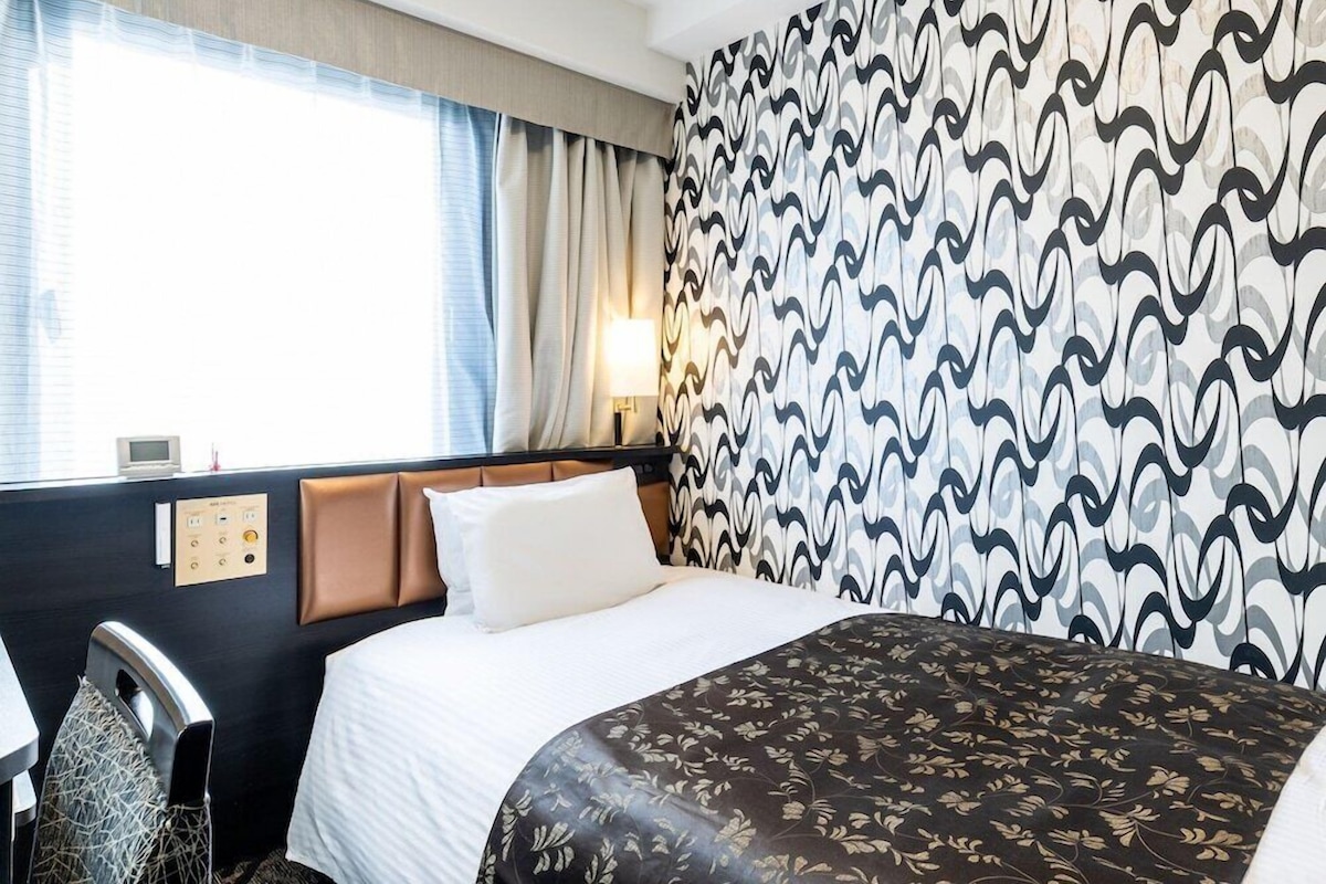 Une chambre d’hôtel avec un lit, un bureau et une chaise. Il y a une fenêtre avec des rideaux, un mur recouvert d’un papier peint à motifs et un panneau de commande fixé au mur.
