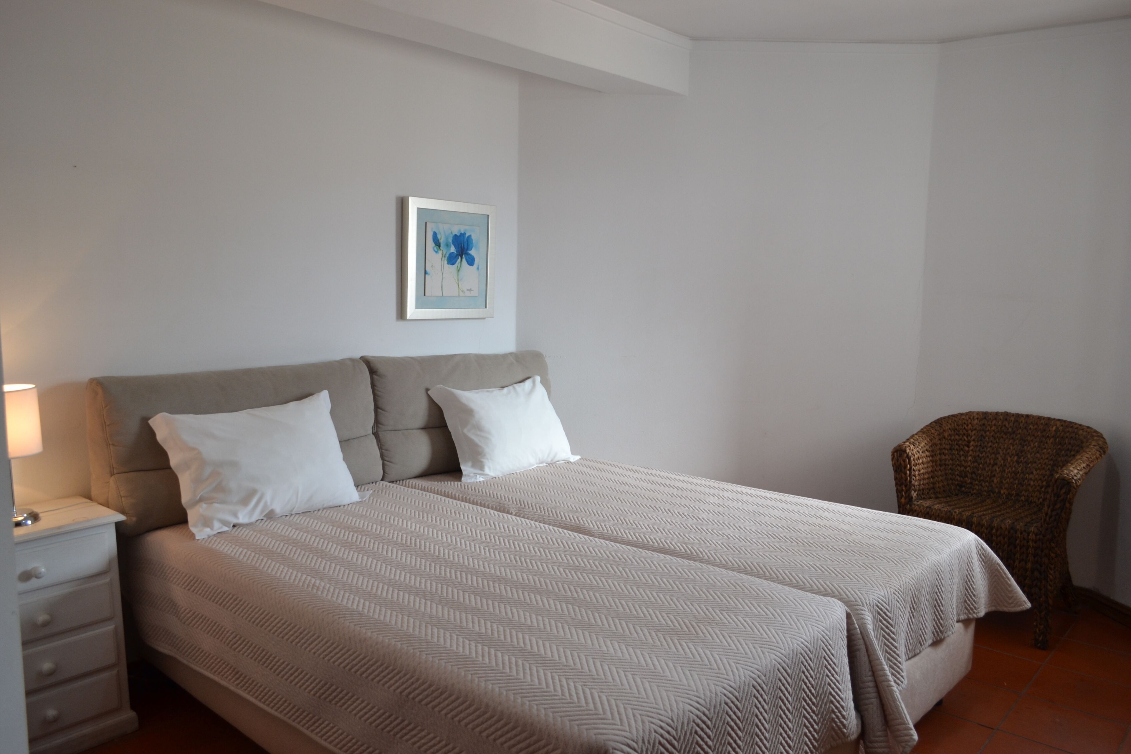 Un lit double avec une couvre-lit à motifs en chevrons, une table de chevet avec une lampe, une chaise en osier et un tableau encadré au mur.