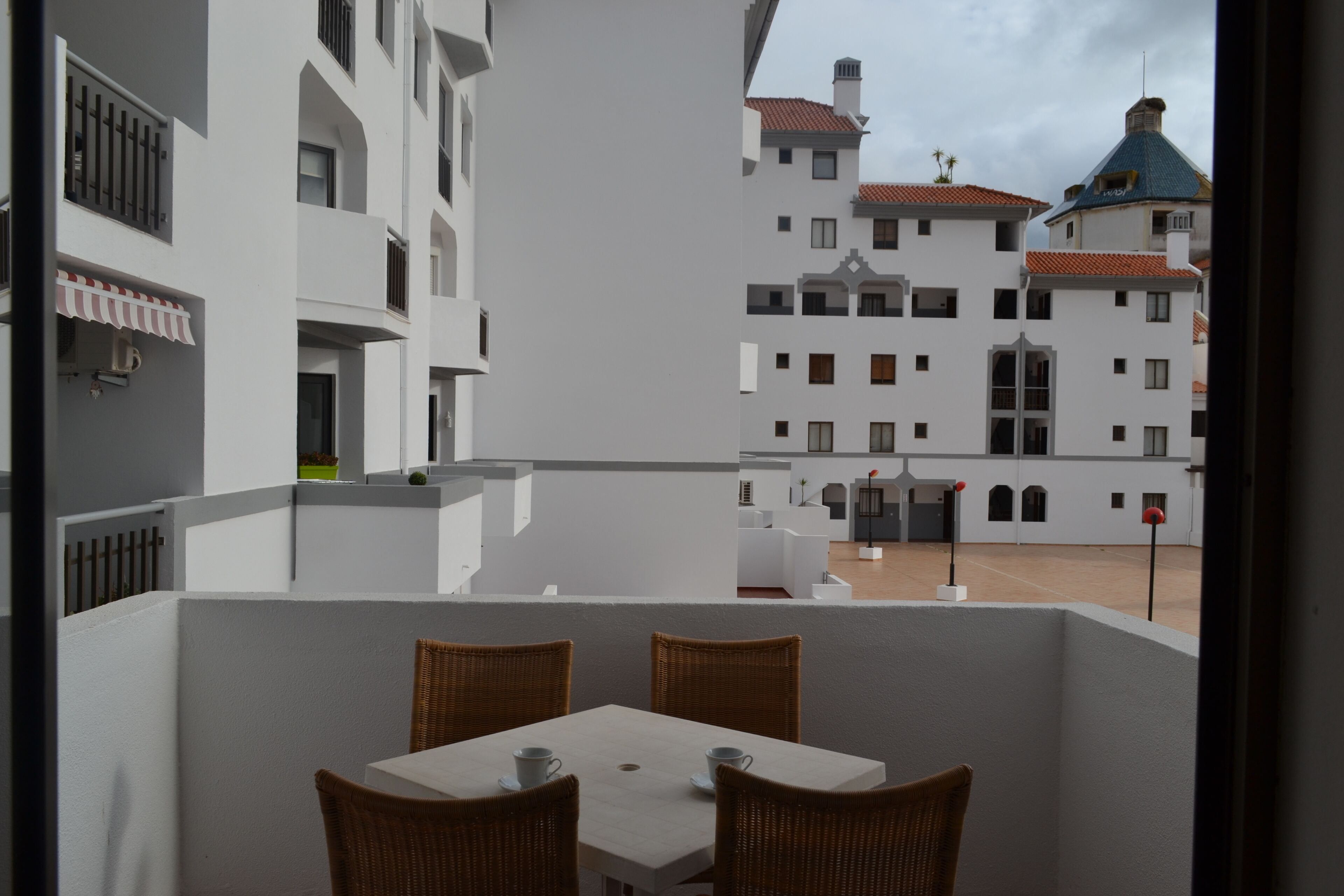 A balcony with wicker chairs and a table, overlooking a courtyard with buildings.
