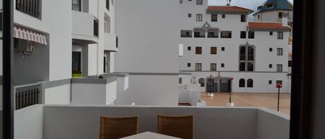 A balcony with wicker chairs and a table, overlooking a courtyard with buildings.