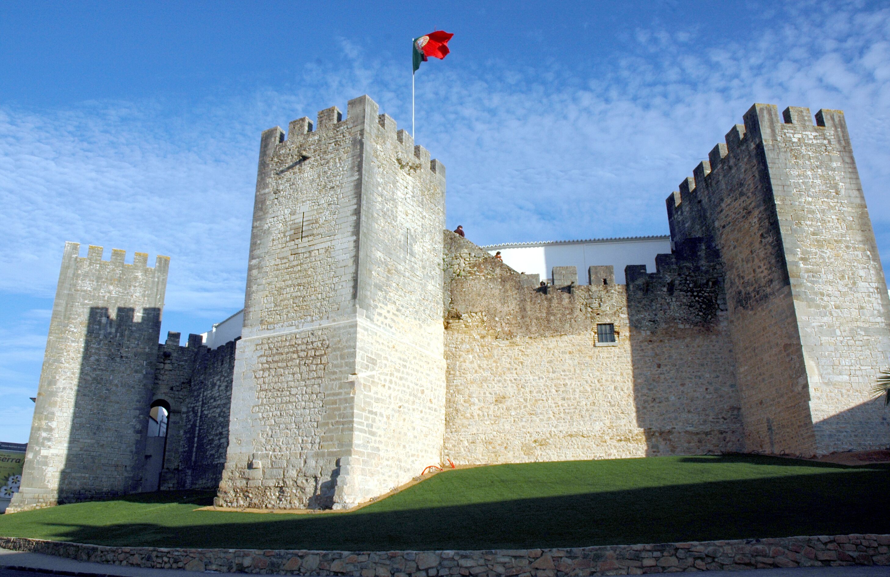 A historic stone castle with two prominent towers, a flagpole with a flag, and a well-maintained lawn in front.
