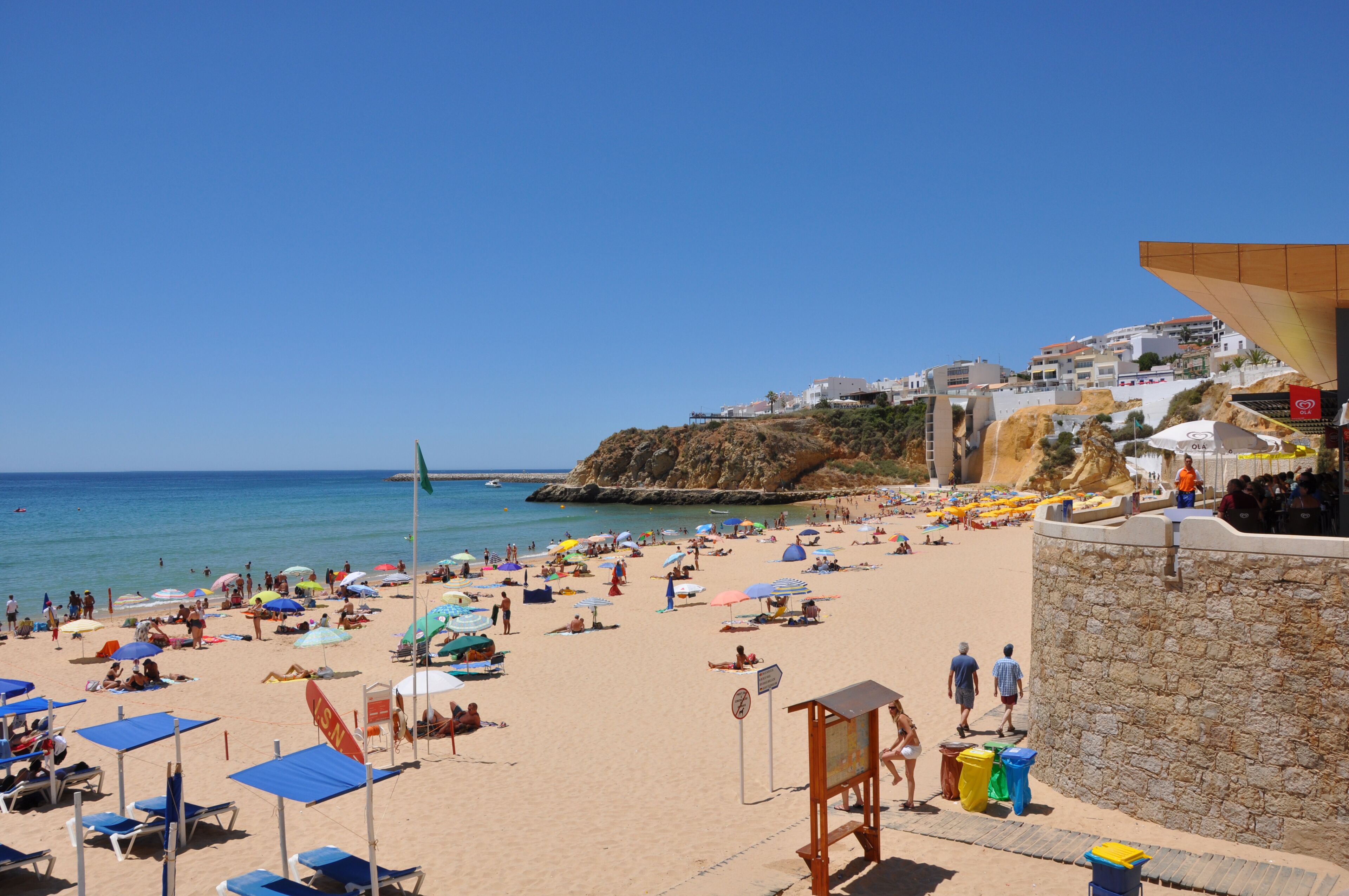 A beach with numerous umbrellas and sun loungers, a lifeguard tower, and a rocky cliff in the background.