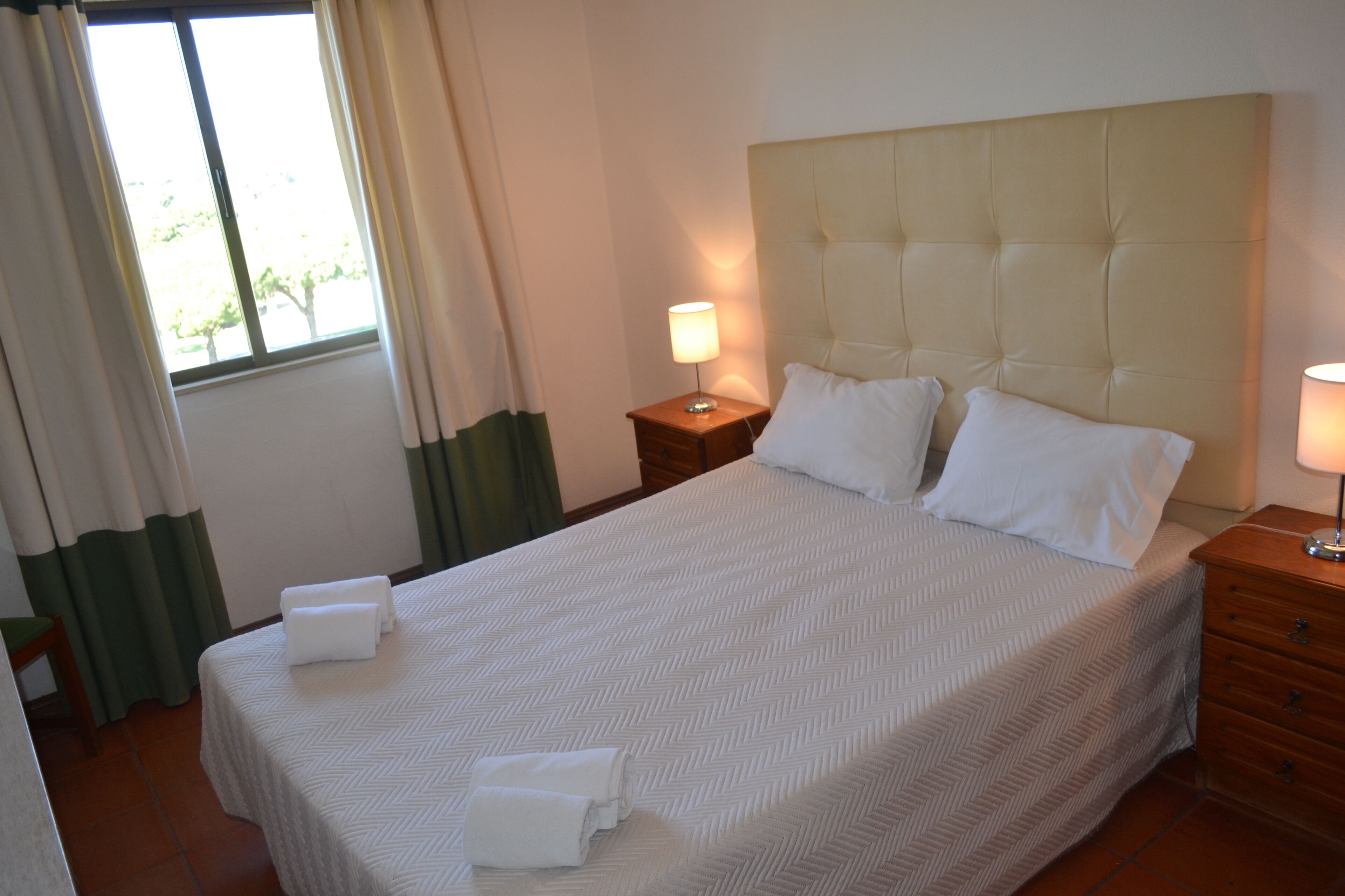 A neatly made bed with white linens and pillows, a bedside table with a lamp, and a window with curtains.