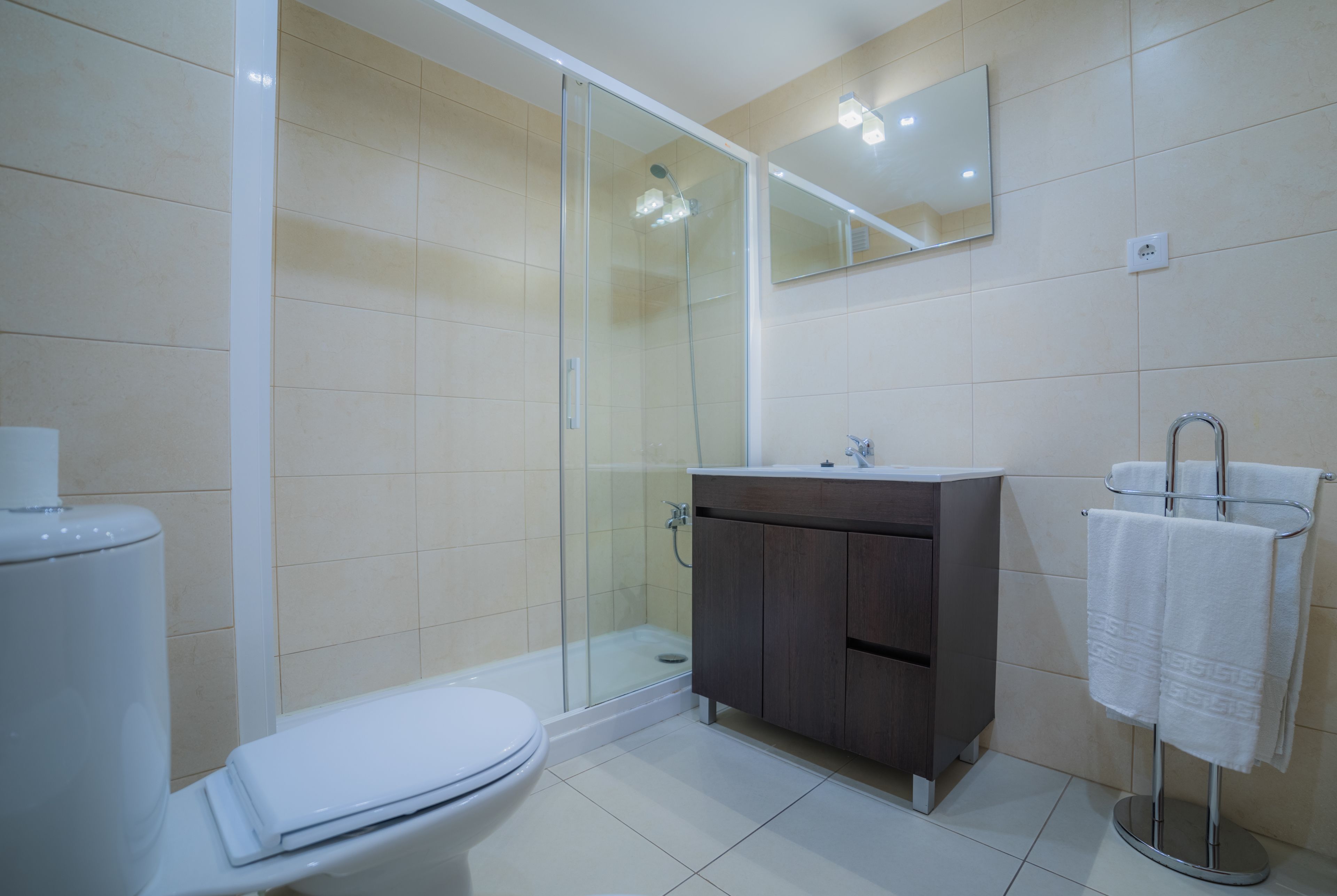 Ein modernes Badezimmer mit einer Glaskabine zur Dusche, einer weißen Toilette, einem dunklen Holzwaschtisch und einem Handtuchhalter mit weißen Handtüchern.