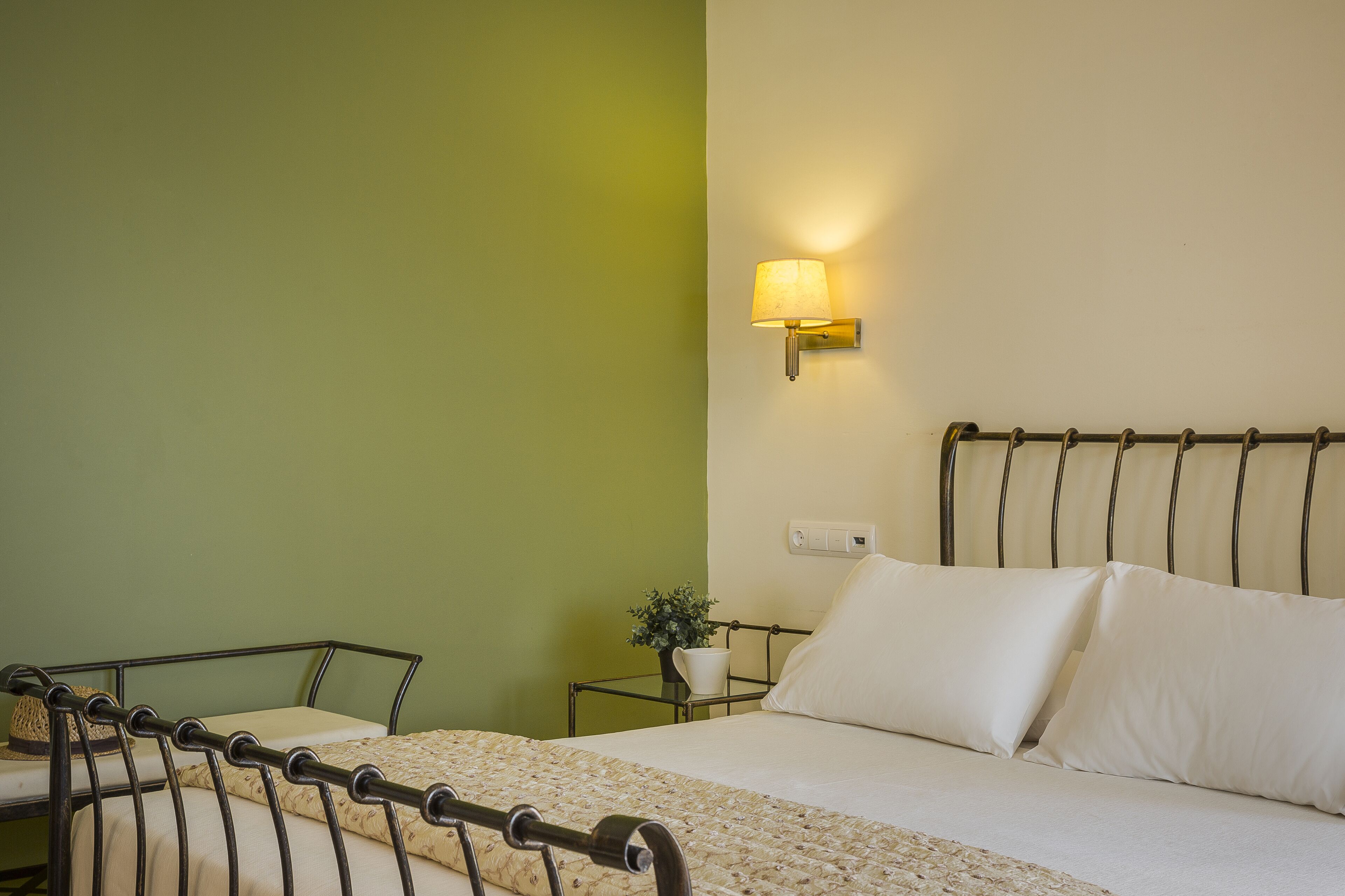 A neatly made bed with white linens and a metal headboard, a bedside table with a lamp, and a small potted plant.