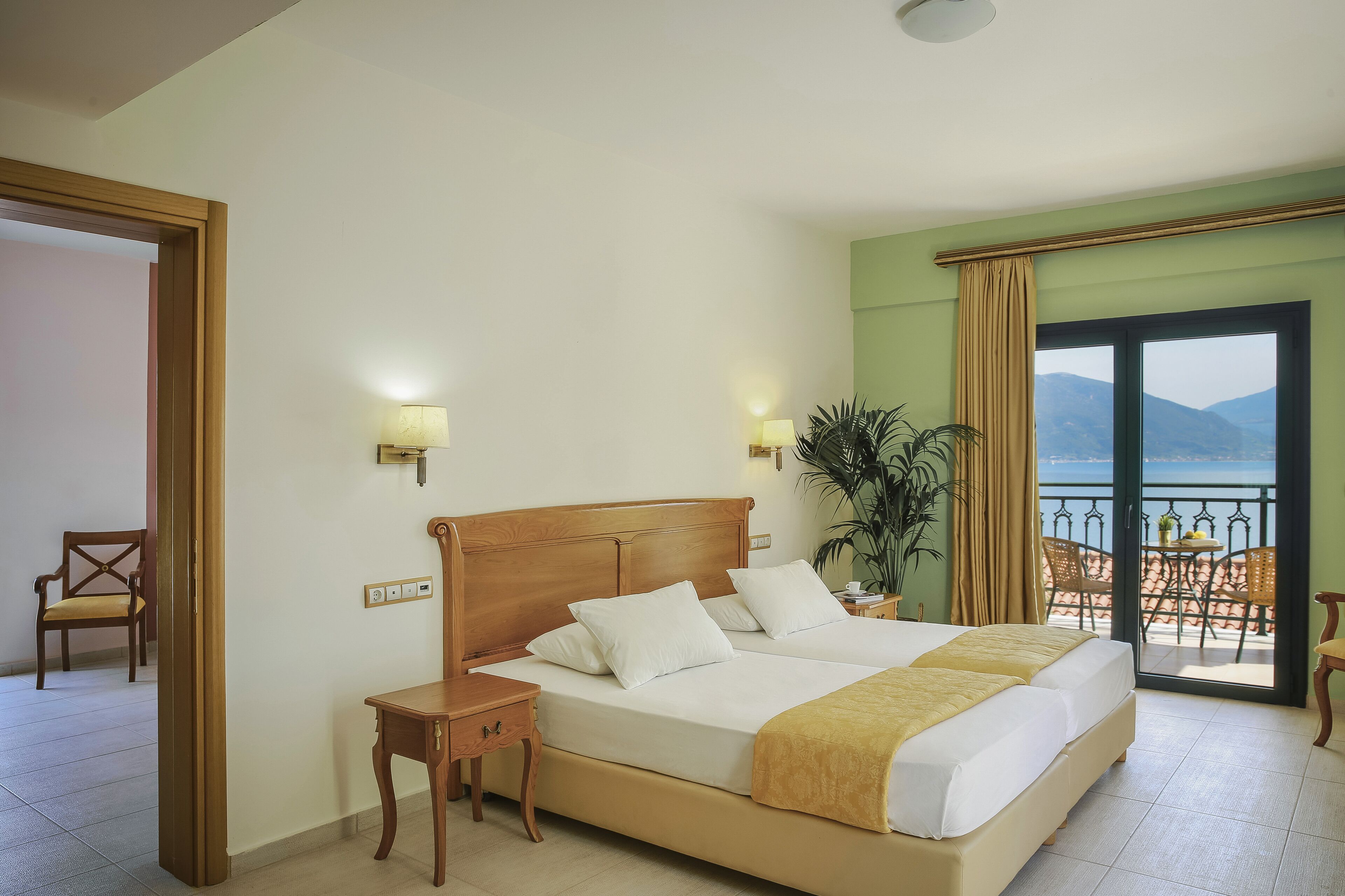 A hotel room with a large bed, a nightstand, a chair, a potted plant, and a balcony with a view of the sea.
