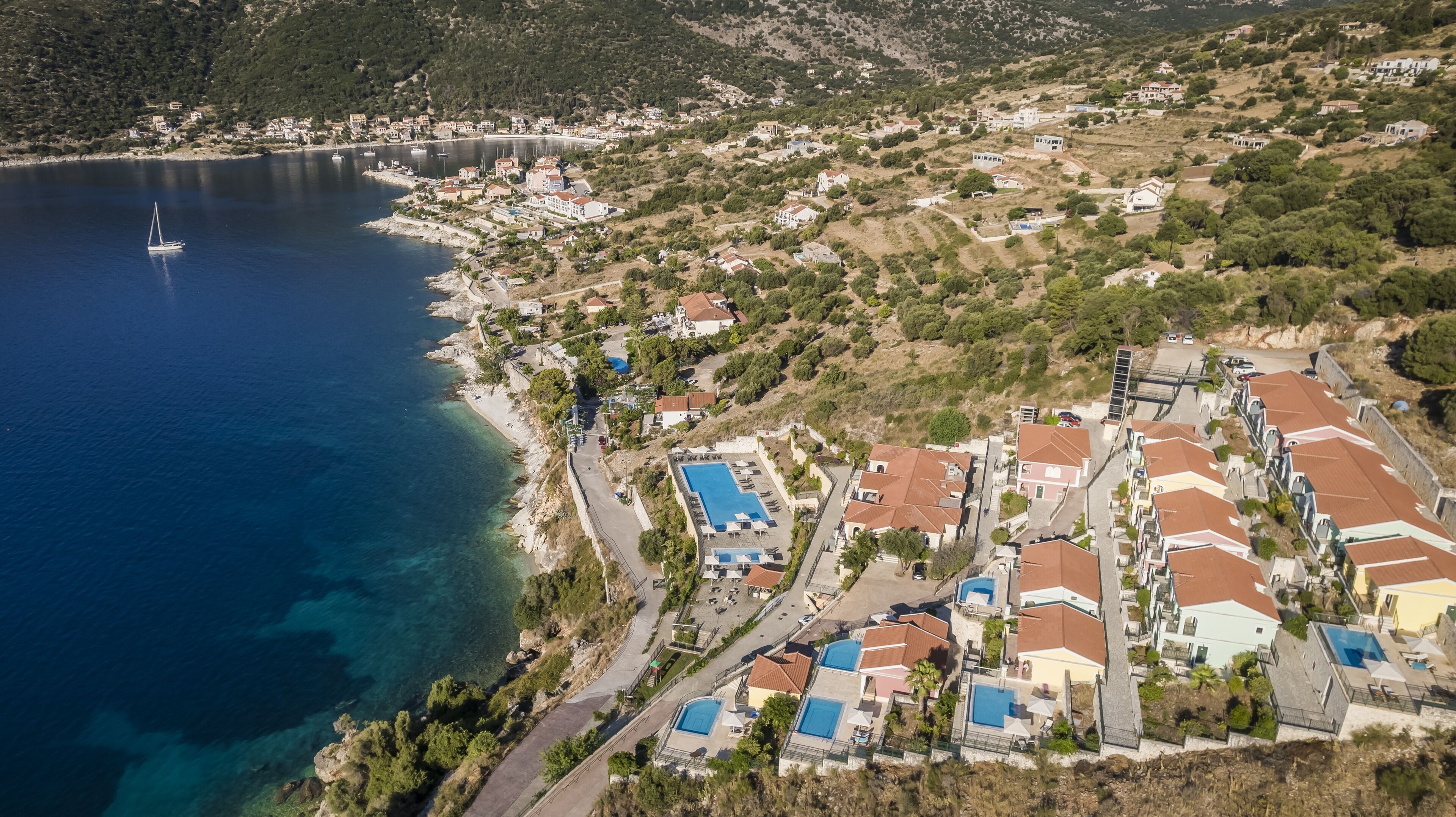 An aerial view of a coastal resort with multiple swimming pools, a marina, and a hillside settlement.
