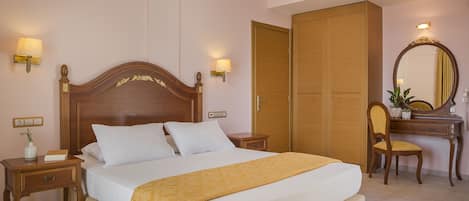 A neatly made bed with a wooden headboard, a bedside table with a lamp, a desk with a chair, and a wardrobe.
