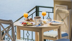 Una mesa con alimentos para el desayuno y un vaso de jugo de naranja, con vista al mar tranquilo y montañas lejanas.