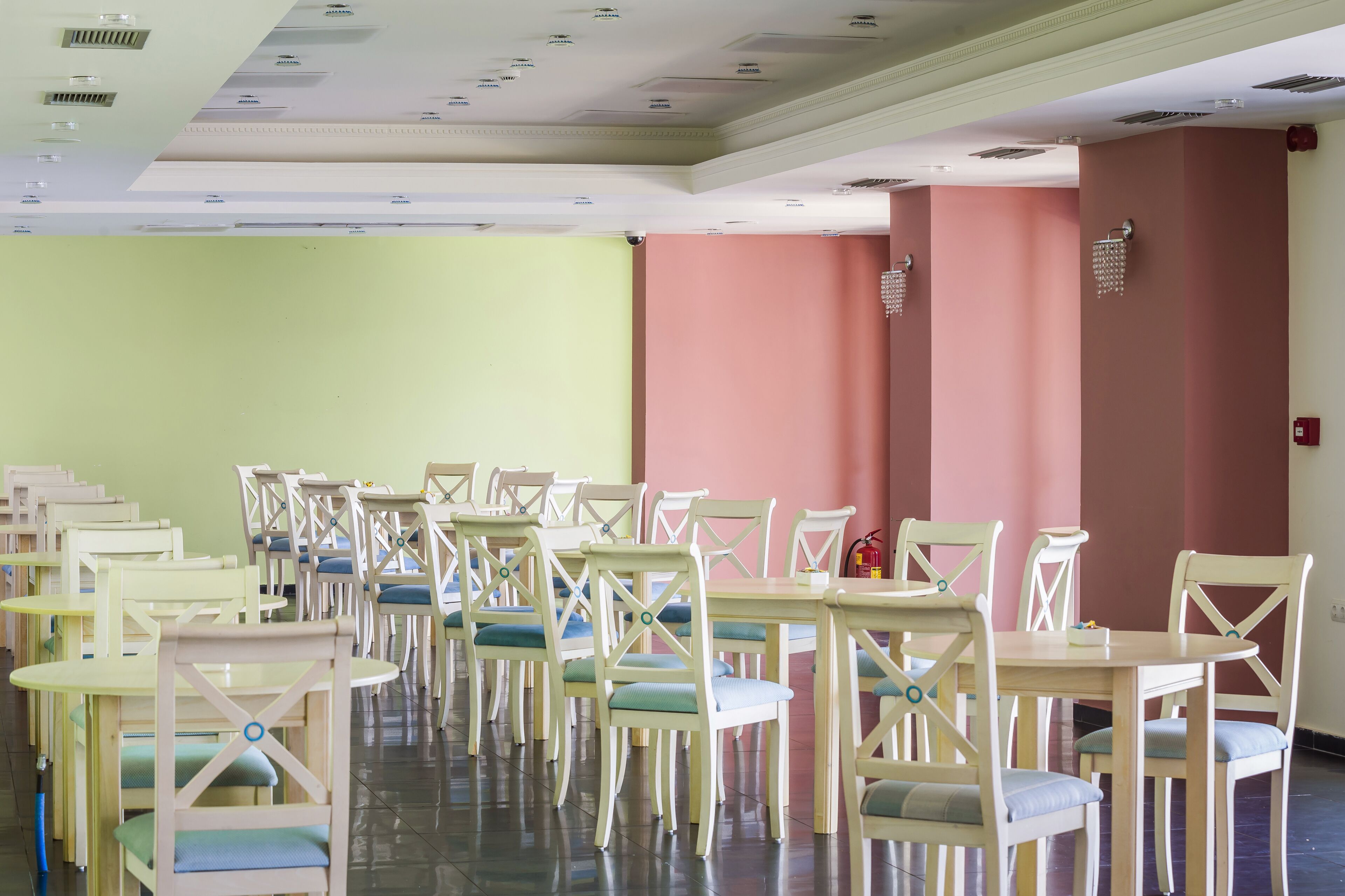 A room with multiple tables and chairs arranged in rows, a fire extinguisher on the wall, and a ceiling with recessed lighting.