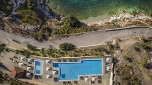 Vista aérea de un complejo turístico con una piscina grande, varias zonas de descanso y un camino de acceso a la playa.