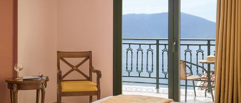 A hotel room with a bed, a chair, a small table with a glass of wine, and a balcony with a view of mountains.