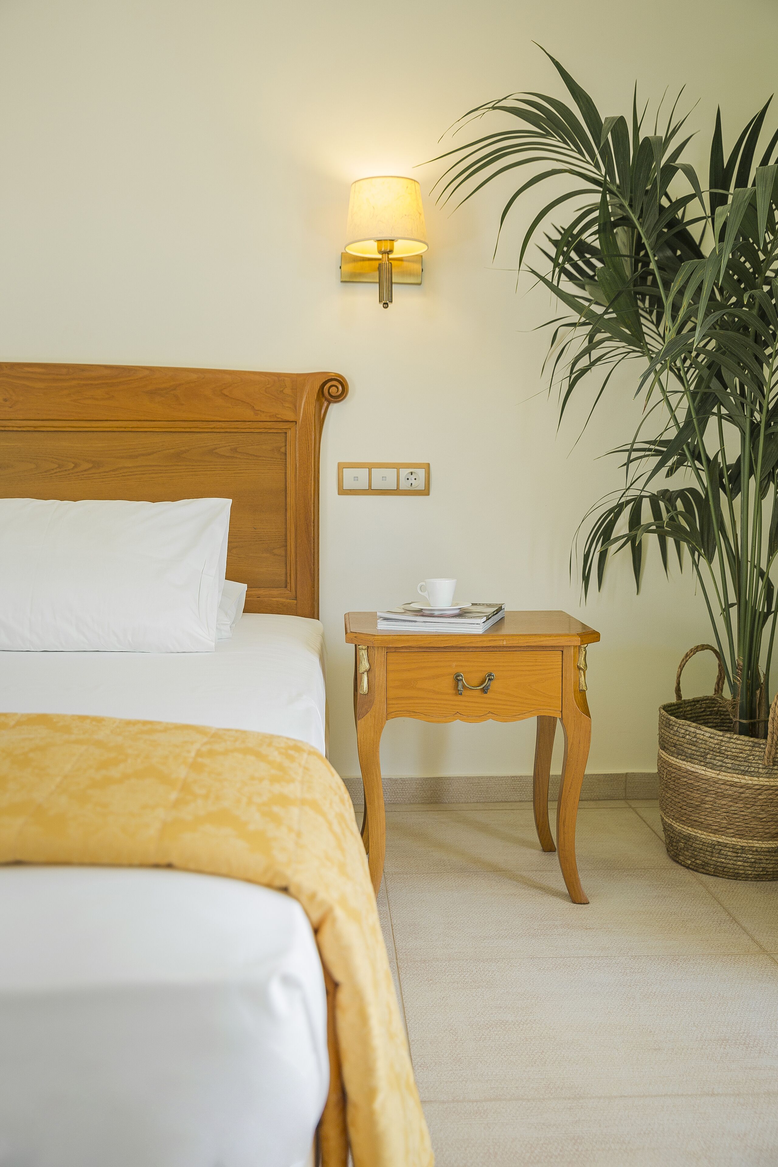 Una cama bien hecha con ropa de cama blanca y amarilla, una mesita de noche con lámpara y una planta en maceta.