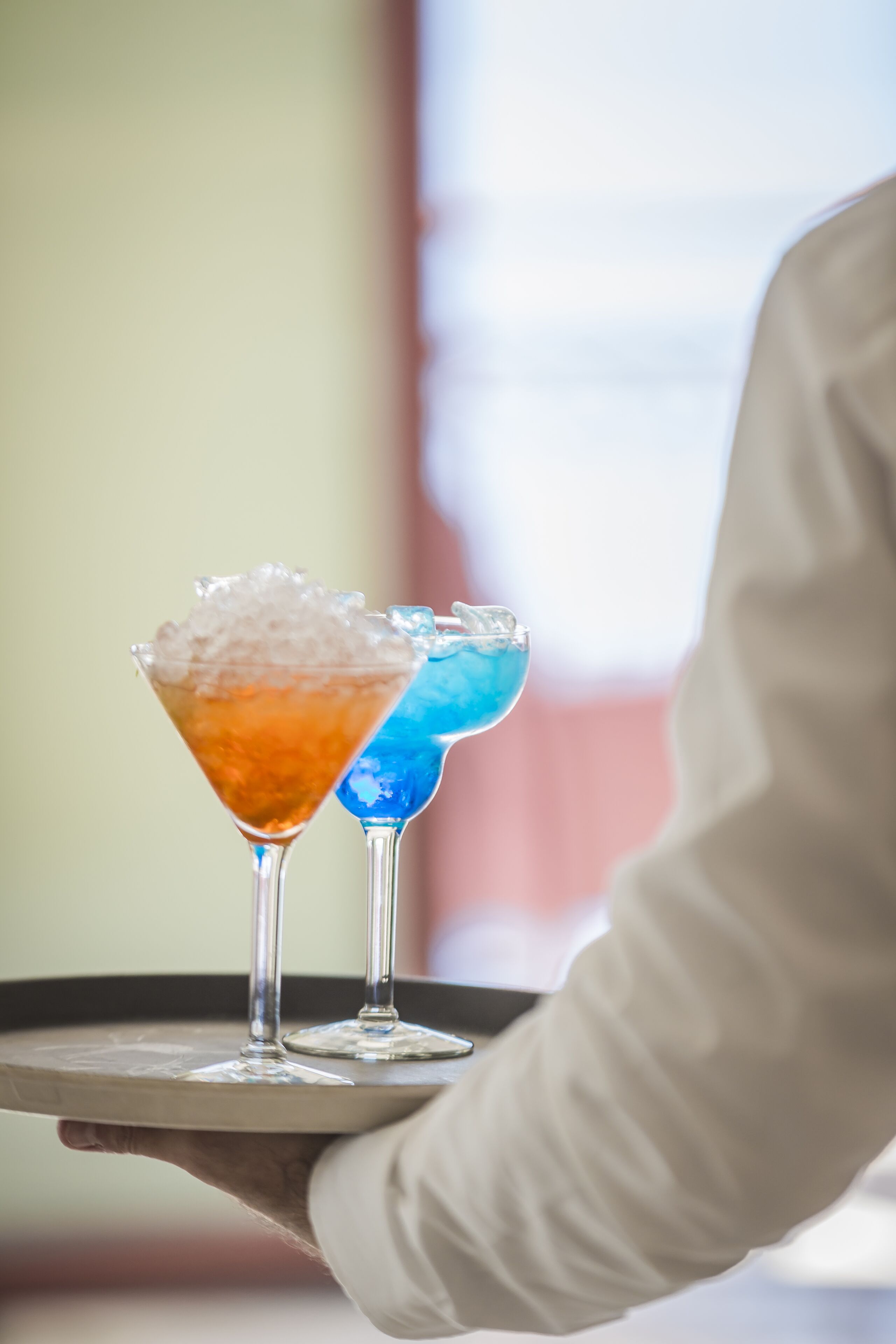 A tray with two martini glasses, one with a clear liquid and the other with a blue liquid, held by a person wearing a white shirt.