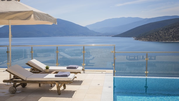Zona junto a la piscina con sillones de descanso y una sombrilla grande, con vista a un cuerpo de agua tranquilo y montañas distantes.