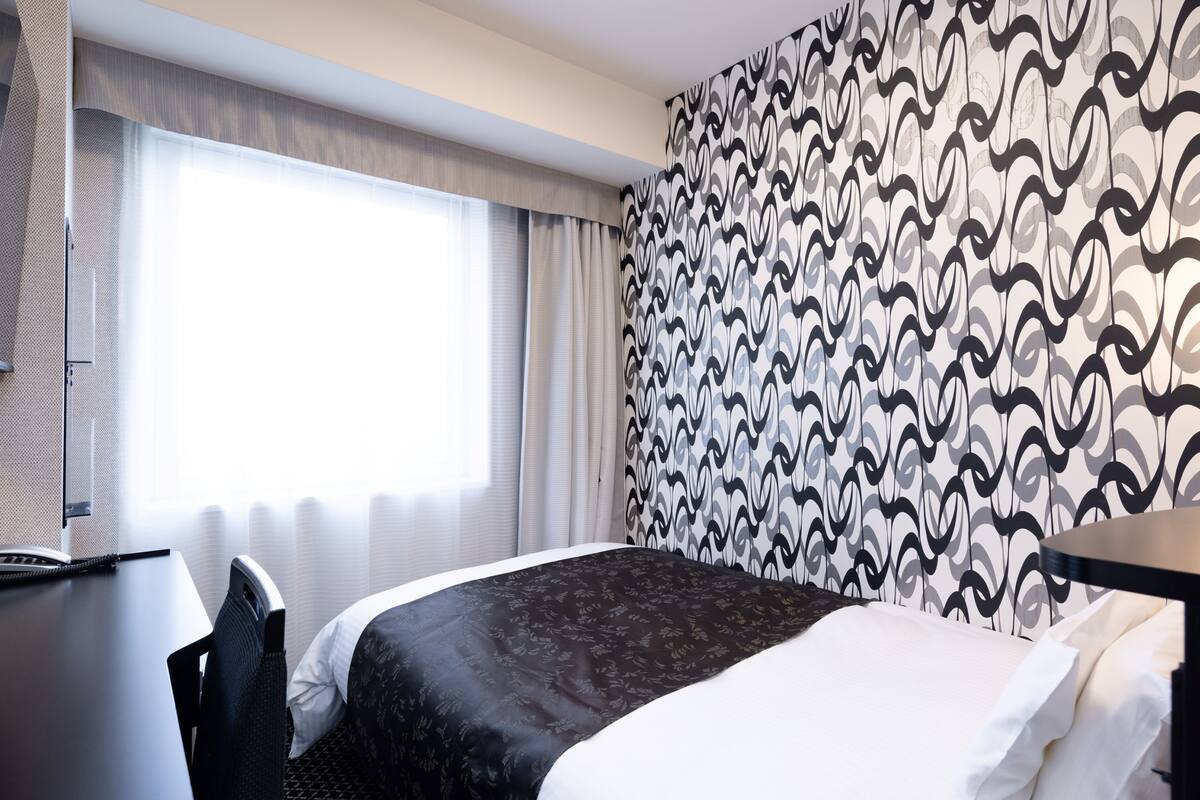 A hotel room with a large bed, a desk, and a chair. There is a window with curtains, and a patterned wall.