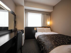 Habitación de hotel con cama, escritorio, silla, mesita, espejo, ventana con cortinas y luz empotrada en la pared.