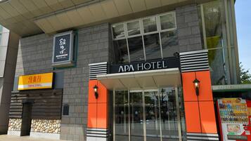 Exterior del APA HOTEL con un diseño moderno, destacando una entrada prominente y una máquina expendedora a la derecha.