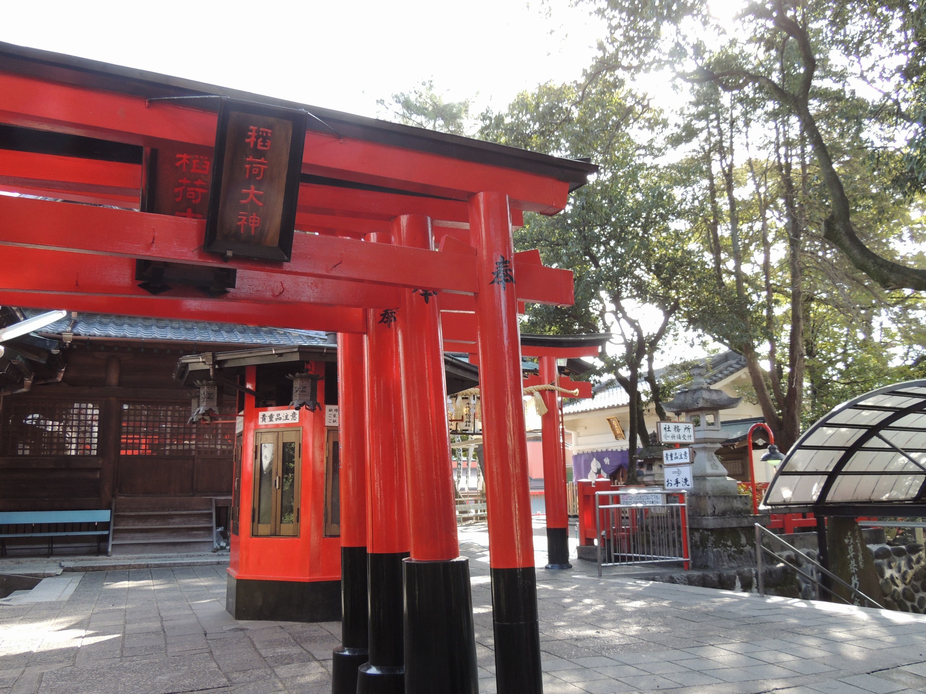 A traditional Japanese torii gate with a red and black color scheme, leading to a building with a tiled roof and a signboard.