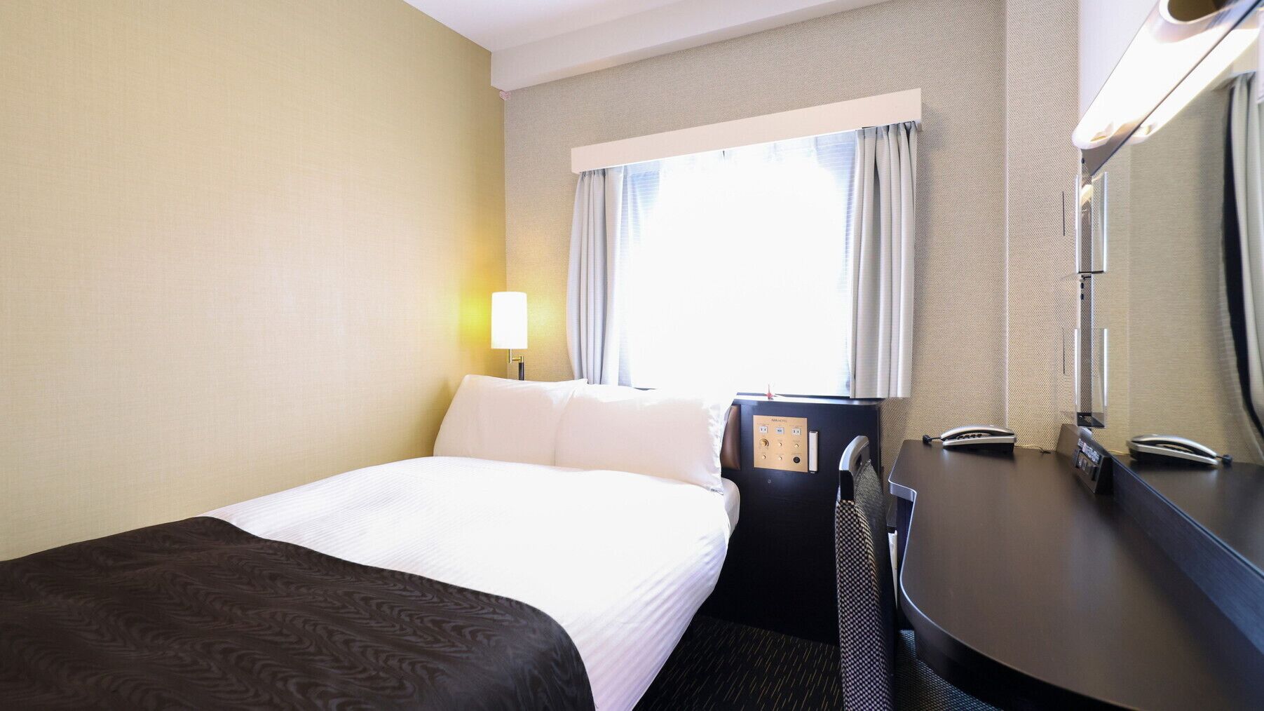 Habitación de hotel con cama, escritorio, silla, lámpara, ventana con cortinas y espejo.