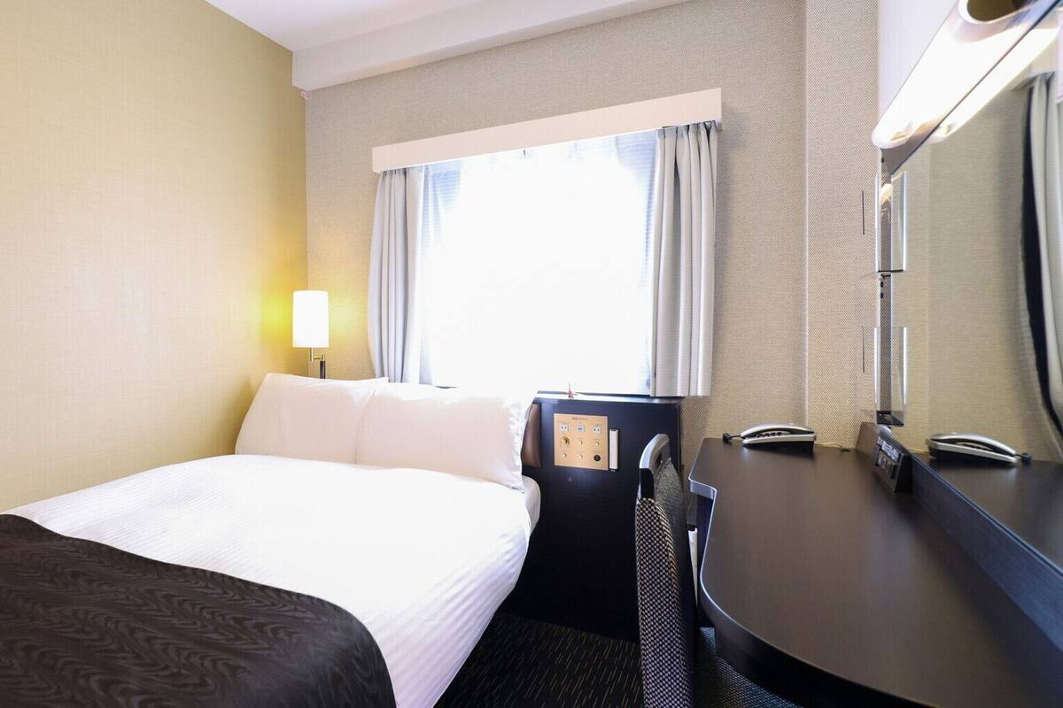 Ett hotellrum med en säng, ett skrivbord, en stol, en lampa, ett fönster med gardiner och en spegel.