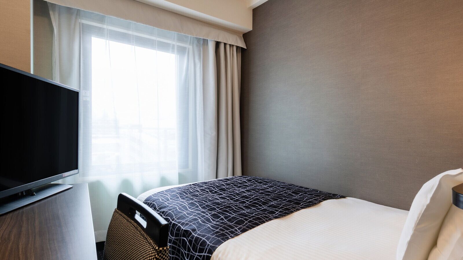 A hotel room with a bed, a television on a stand, a window with curtains, and a plain wall.