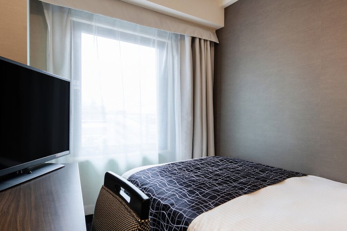 Ett hotellrum med en säng, en tv på ett stativ, ett fönster med gardiner och en enkel vägg.