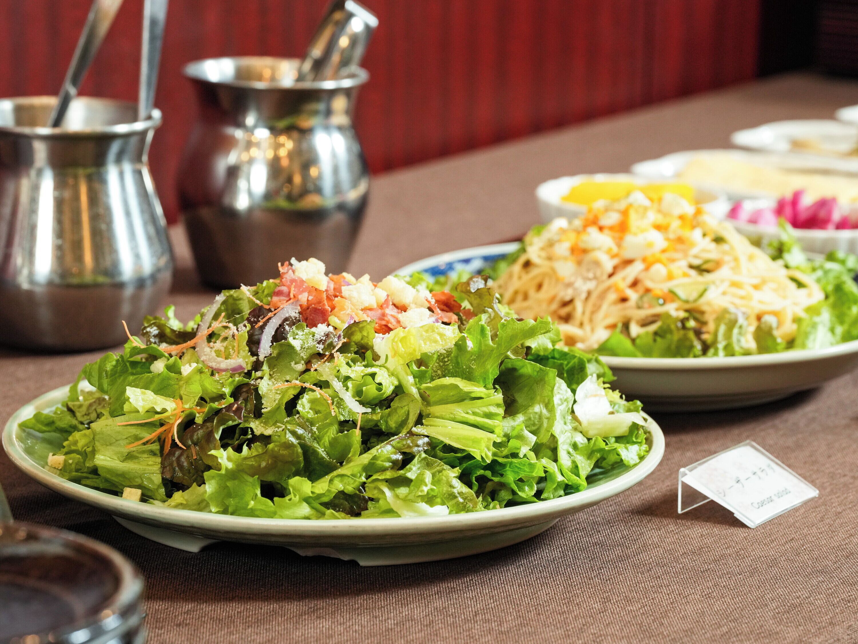 A table with various dishes, including a salad with mixed greens and toppings, a noodle dish with shredded cheese, and a bowl of soup. There are also two metal pitchers and a small card on the table.