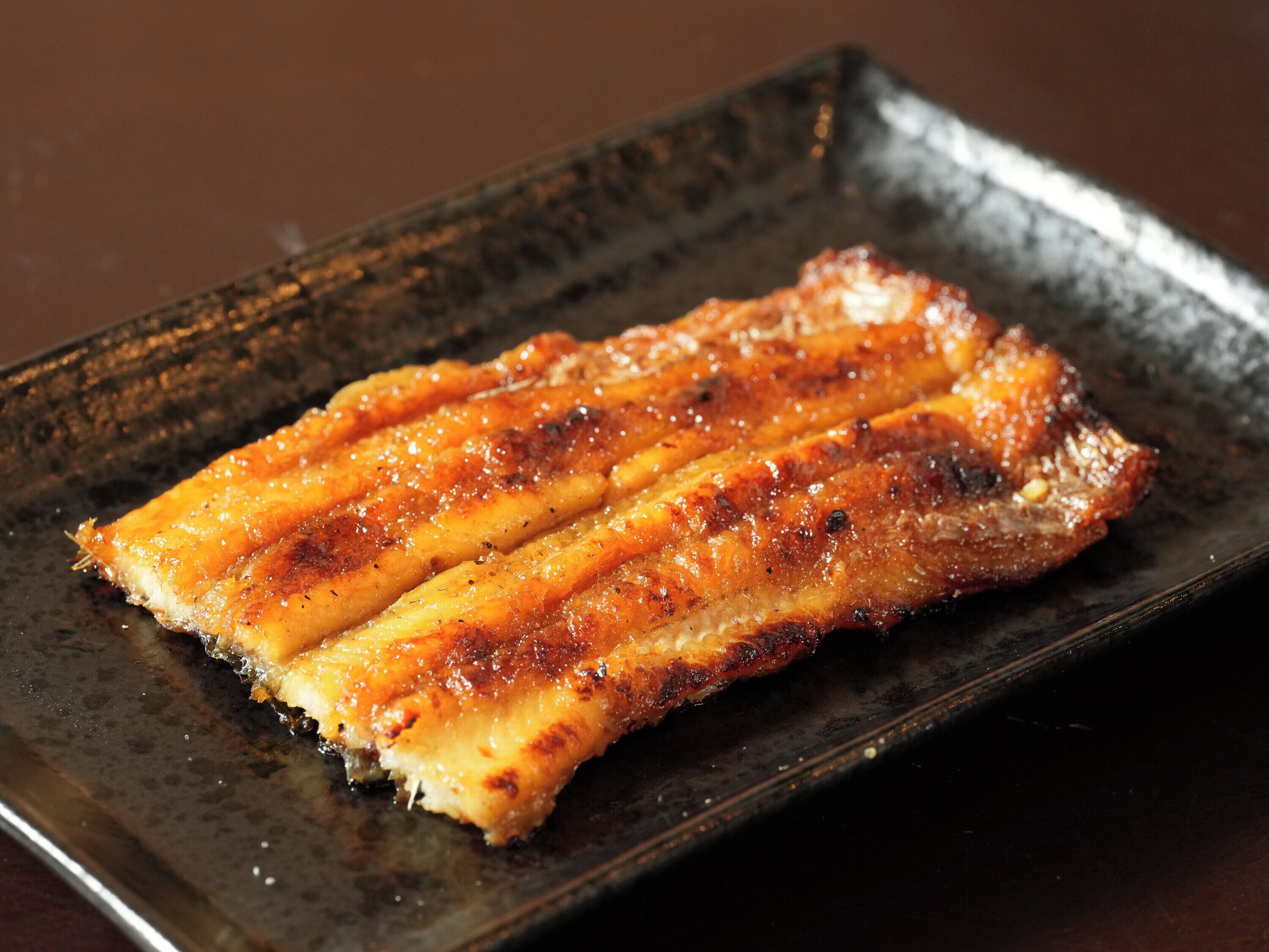 A rectangular plate with a grilled dish, possibly pork belly, on a dark, textured surface.