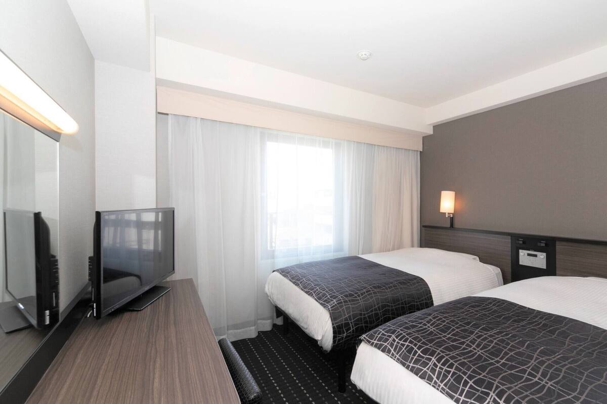 A hotel room with two beds, a TV, a desk, and a window with curtains.