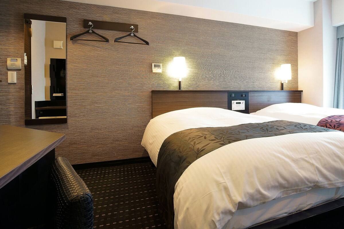 A hotel room with two beds, a desk, a mirror, and a wall-mounted light fixture.