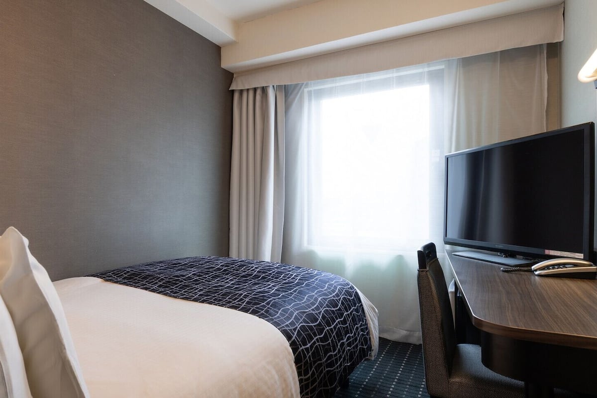 Ett hotellrum med en säng, ett skrivbord, en tv och ett fönster med gardiner.