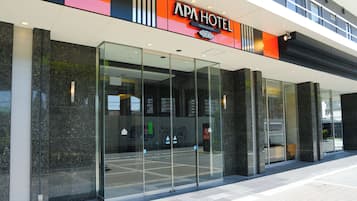 Une entrée d’hôtel moderne avec une façade en verre, une enseigne bien visible indiquant « APA HOTEL », et un espace de stationnement clairement délimité, avec un panneau indiquant les places réservées aux personnes handicapées.