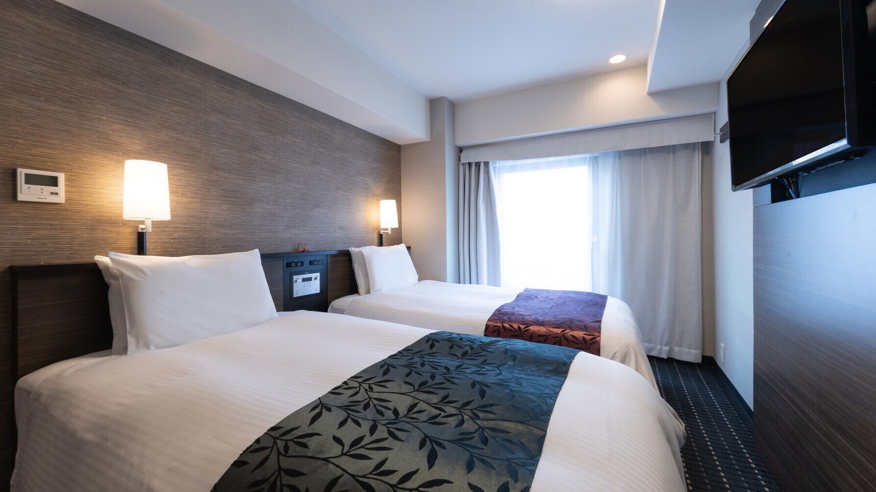 A hotel room with two beds, a TV, a thermostat, and a window with curtains.