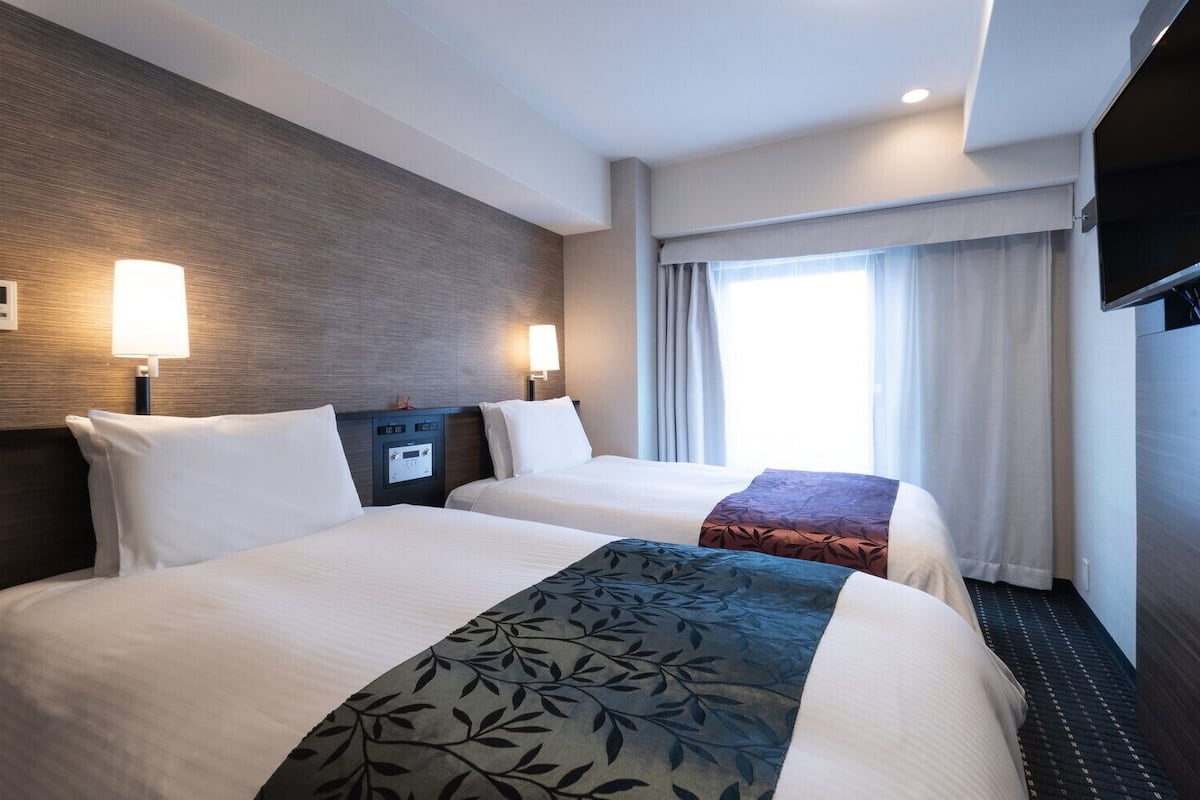 Ett hotellrum med två sängar, en TV, en termostat och ett fönster med gardiner.