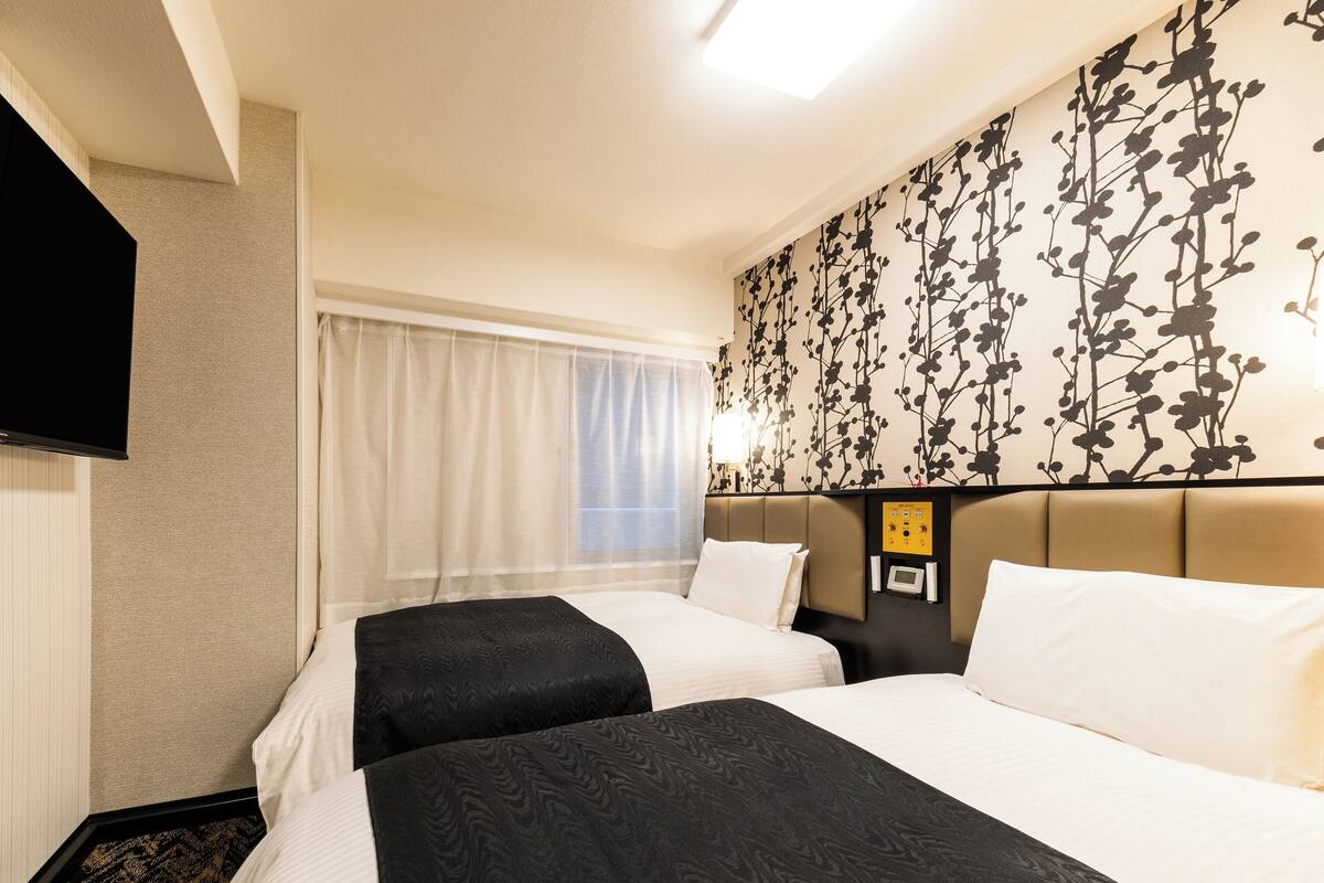 Habitación de hotel con dos camas, un televisor de pantalla plana, panel de control en la pared y una ventana con cortinas translúcidas.