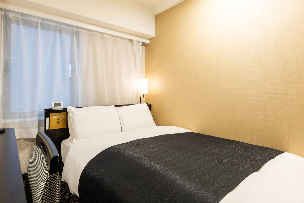 Una habitación de hotel con una cama, una mesita de noche, una lámpara y una ventana con cortinas.