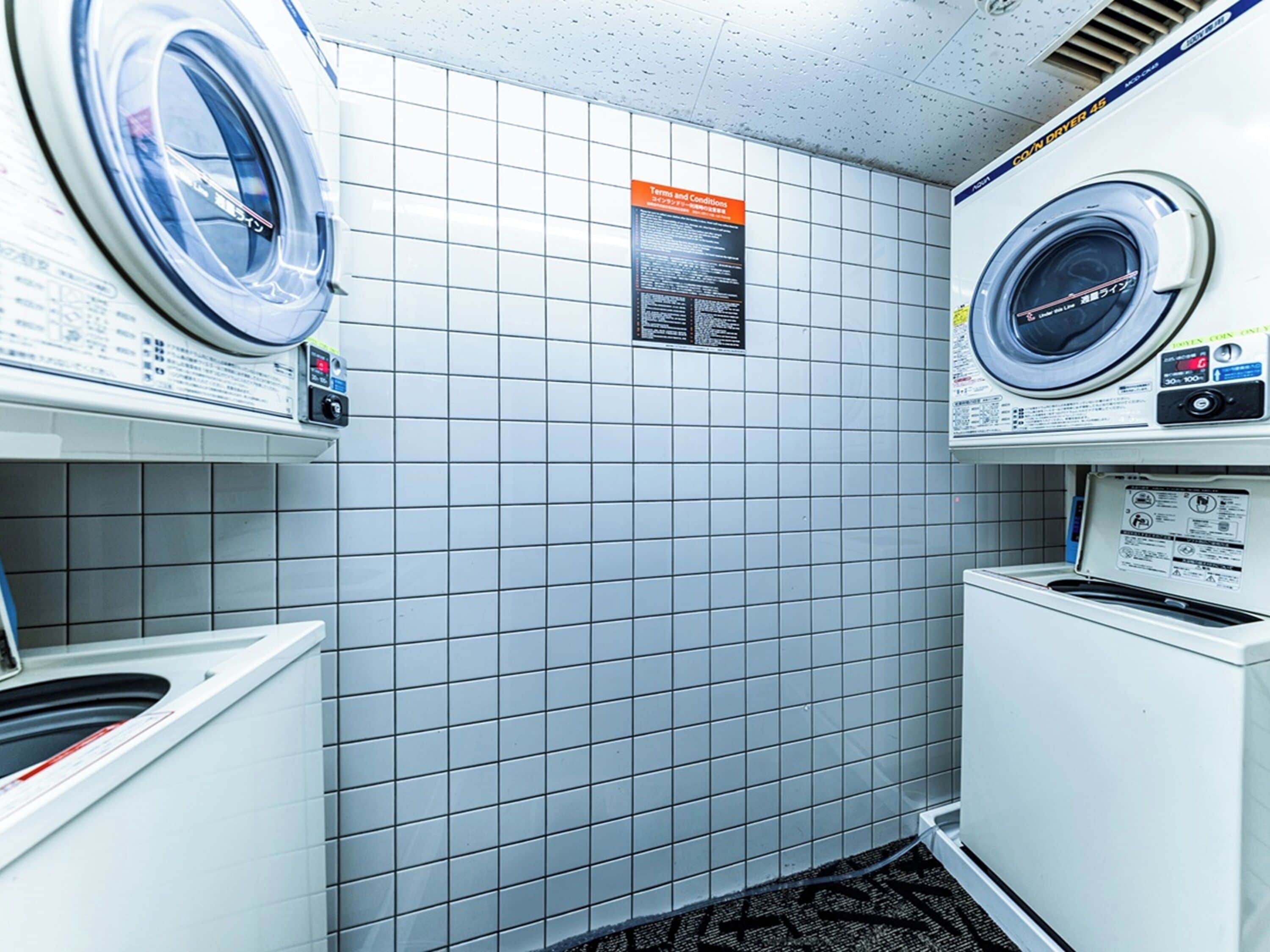 A laundry room with two washing machines, a dryer, and a tiled wall with a terms and conditions sign.