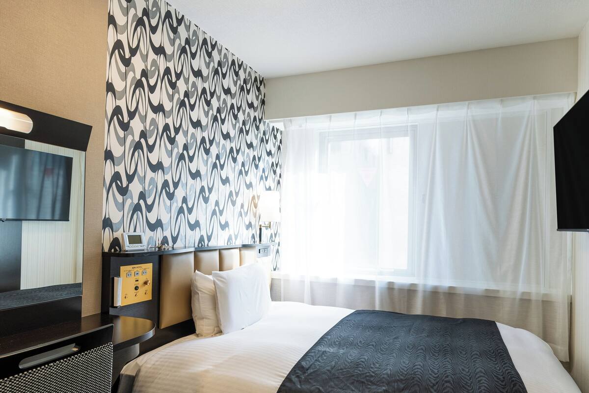 Habitación de hotel con una cama grande, un televisor de pantalla plana, una mesita de noche con lámpara y un papel tapiz estampado.