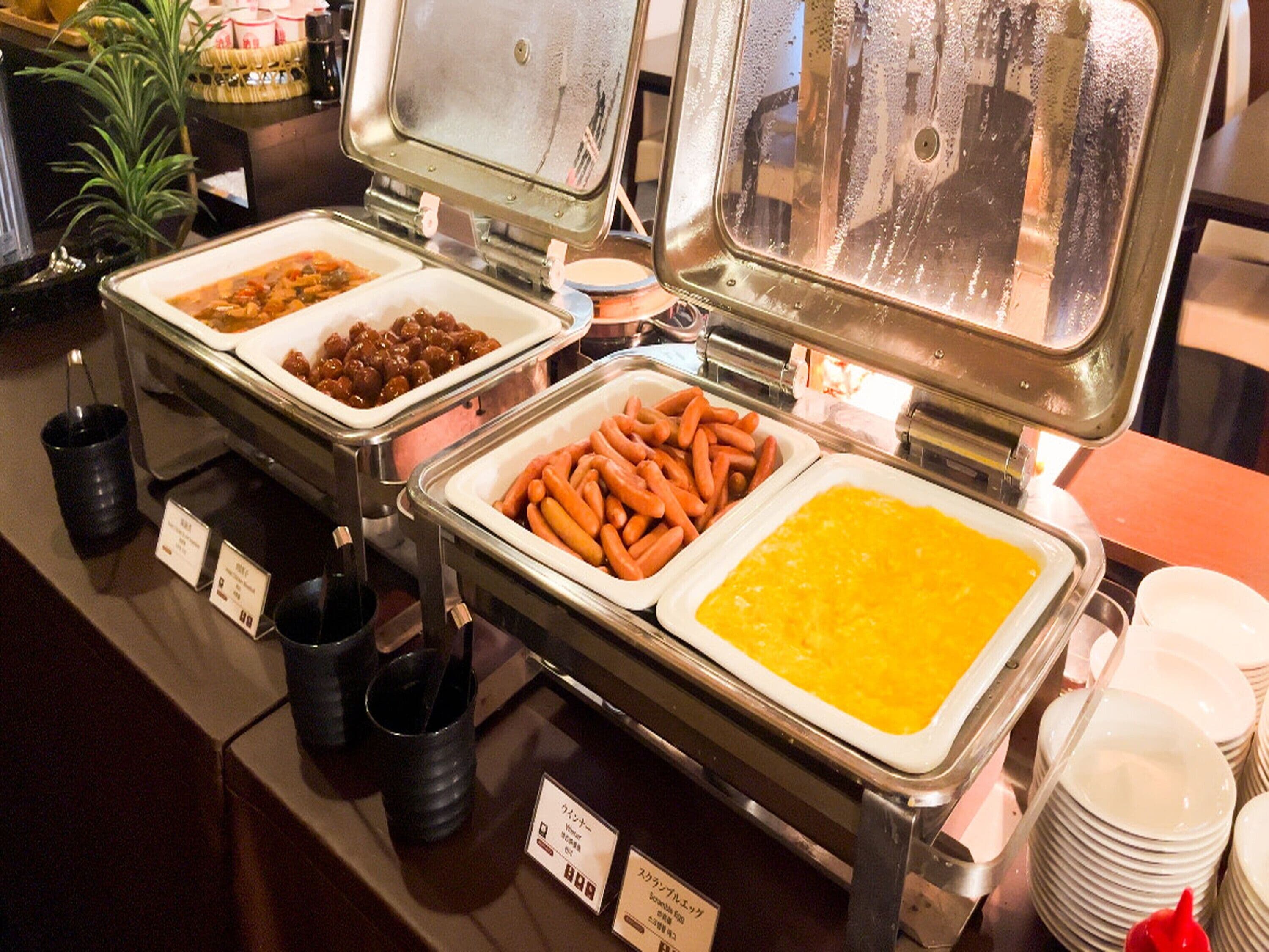 Un bufé con distintos alimentos, incluyendo perros calientes, un recipiente amarillo y un contenedor que parece ensalada o salsa.