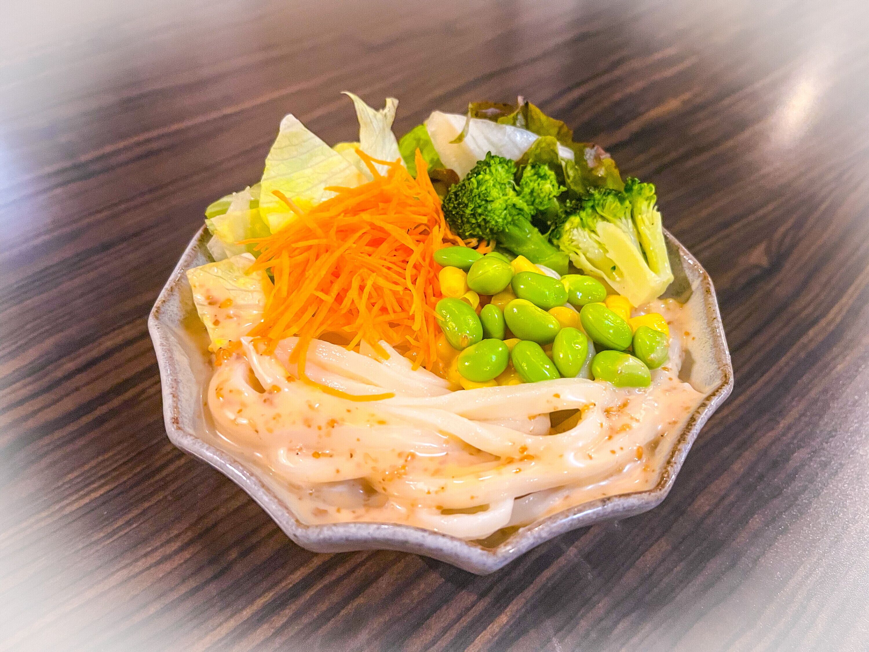 A bowl of ramen with various toppings including noodles, shredded carrots, broccoli, and edamame.